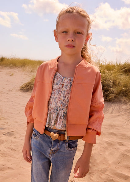 Young girl in a peach jacket and blue jeans standing on a sandy beach with grass and sky in the background.