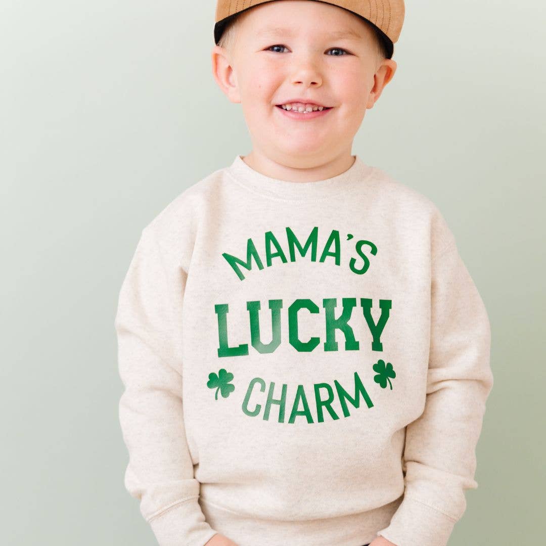 Child wearing a cream sweatshirt with 'Mama's Lucky Charm' text and green shamrock graphics on a light gray background.