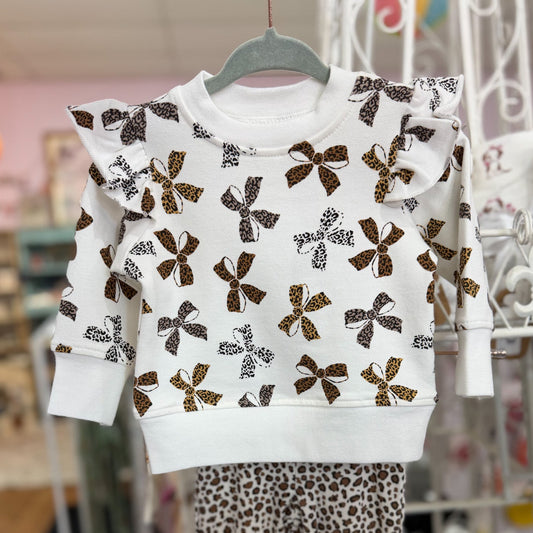 A toddler girl's two-piece outfit hanging on a hanger, featuring a white long-sleeve top with ruffle shoulders and brown bow patterns, paired with leopard print pants.
