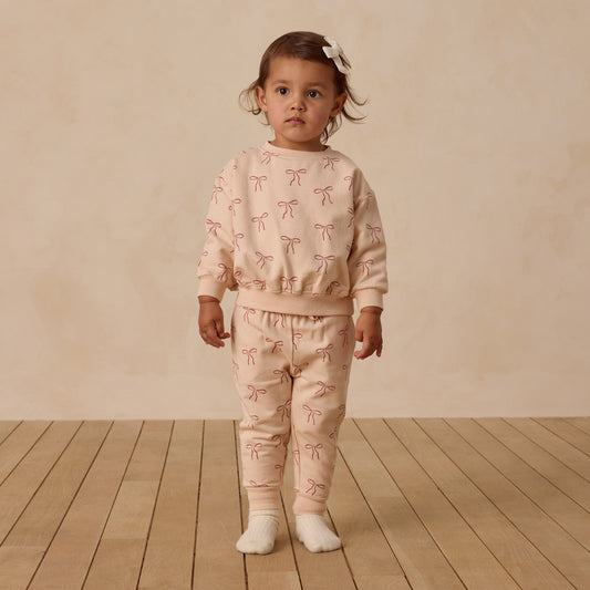Child wearing a pink outfit with bow patterns on a wooden floor and beige wall background