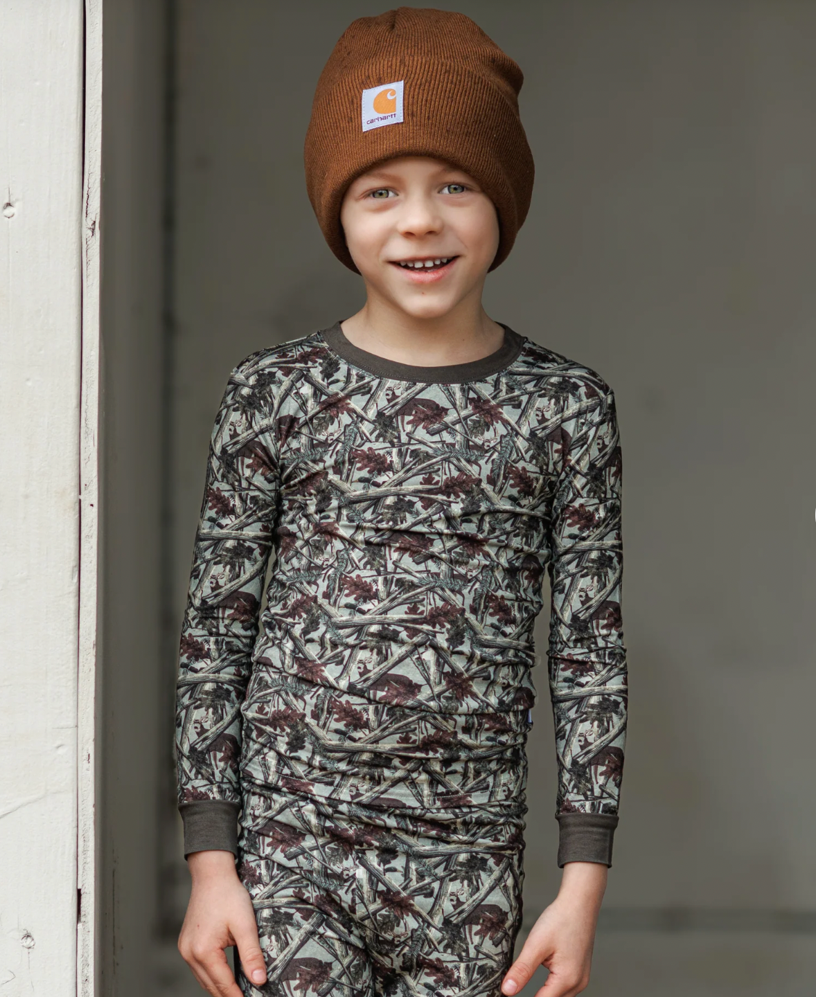 Child wearing a camouflage long-sleeve shirt and pants with a brown beanie, standing against a neutral background.