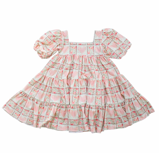 Puff sleeve dress on white background, dress has a block print reminiscent of quilting with faint watercolor looking pink hearts in the center on some blocks, and a floral print in other blocks. The skirt of the dress is sewn into three separate tiers, and the waist is in the empire style with a square neckline.