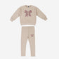 A two-piece children's ribbed knit pajama set in beige, featuring a long-sleeve top with a large multicolor bow graphic on the chest and matching pants with a smaller bow graphic on the upper thigh.