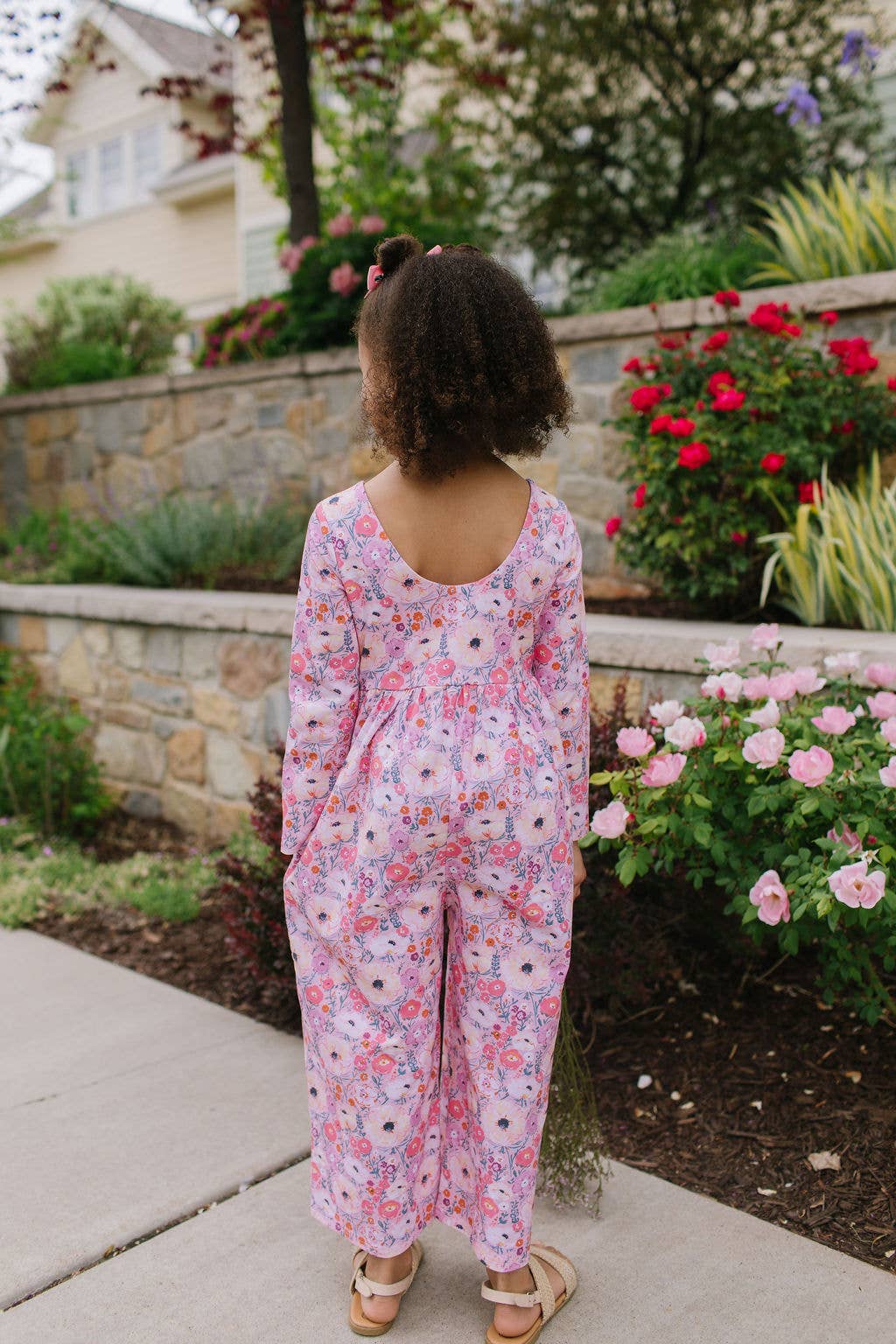 Person wearing a pink floral jumpsuit standing in a garden with flowers and a stone wall.