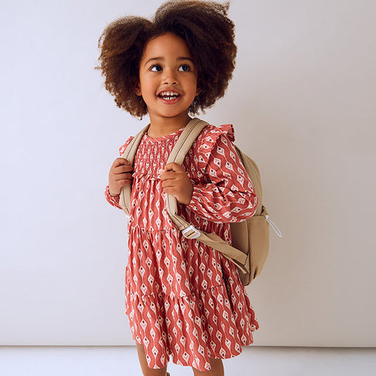 Child wearing a red patterned dress and brown boots with a beige backpack on a white background