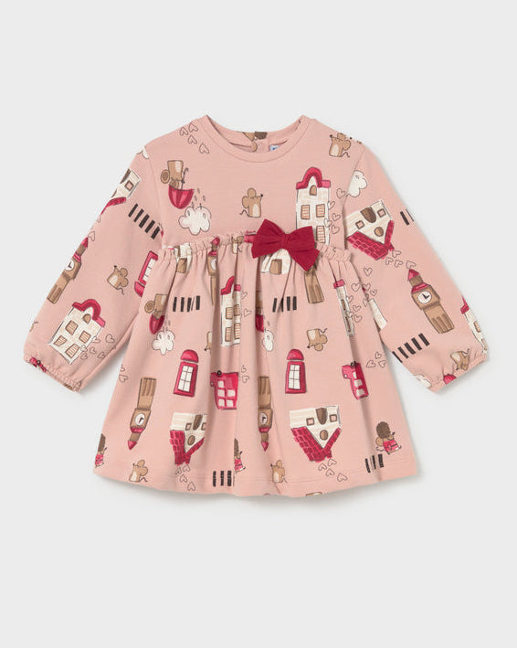 A long-sleeve baby dress in dusty pink with an all-over London-themed print of red buses, tea time phones, and houses, featuring a dark red bow at the waist.