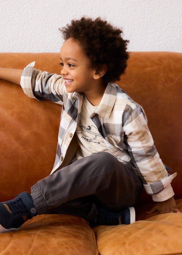 Child sitting on a brown couch wearing a plaid shirt and gray pants.