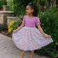 Young girl in a pink and floral dress standing outdoors with greenery in the background