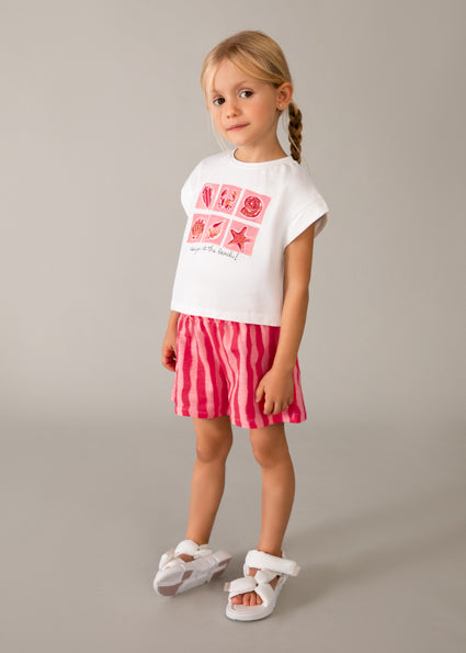 Young girl wearing a white t-shirt with pink graphics and red striped shorts on a gray background