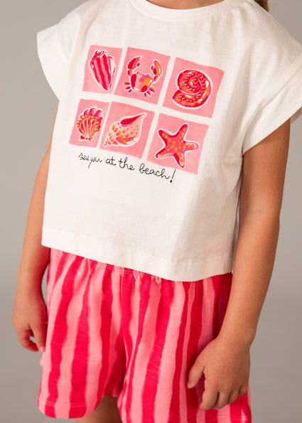Child wearing a white t-shirt with pink sea-themed graphics and red striped shorts on a gray background