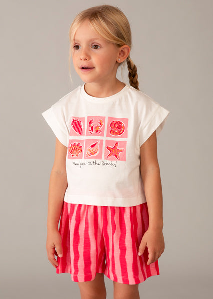 Young girl wearing a white t-shirt with pink graphics and red striped shorts on a gray background