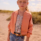 Young girl in a peach jacket and blue jeans standing on a sandy beach with grass and sky in the background.