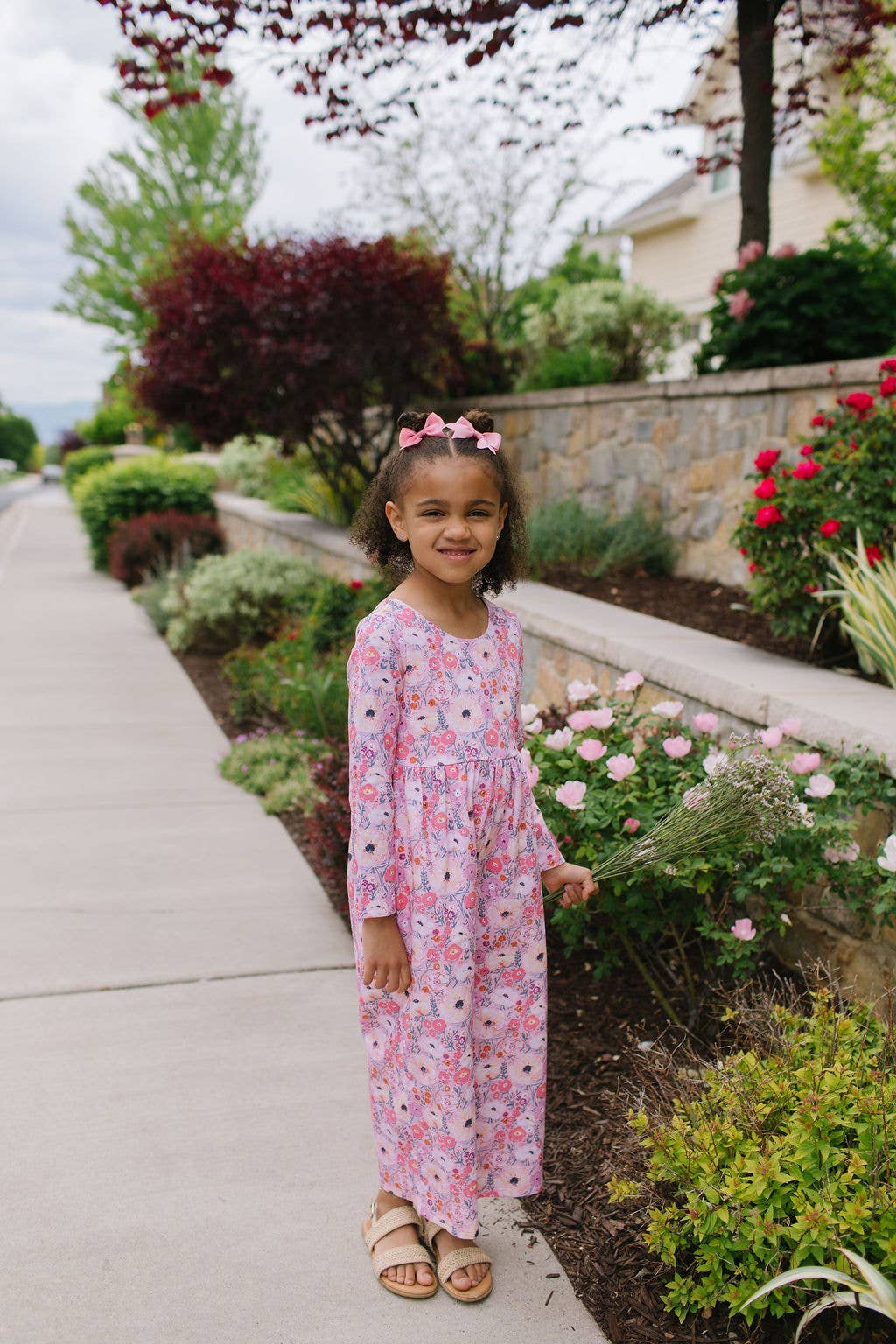 Young girl in a floral dress standing on a sidewalk with garden and house in the background