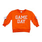 An orange crewneck sweatshirt with 'GAME DAY' printed in white block letters on the front, featuring white bow details on the sleeves, shown against a plain white background.