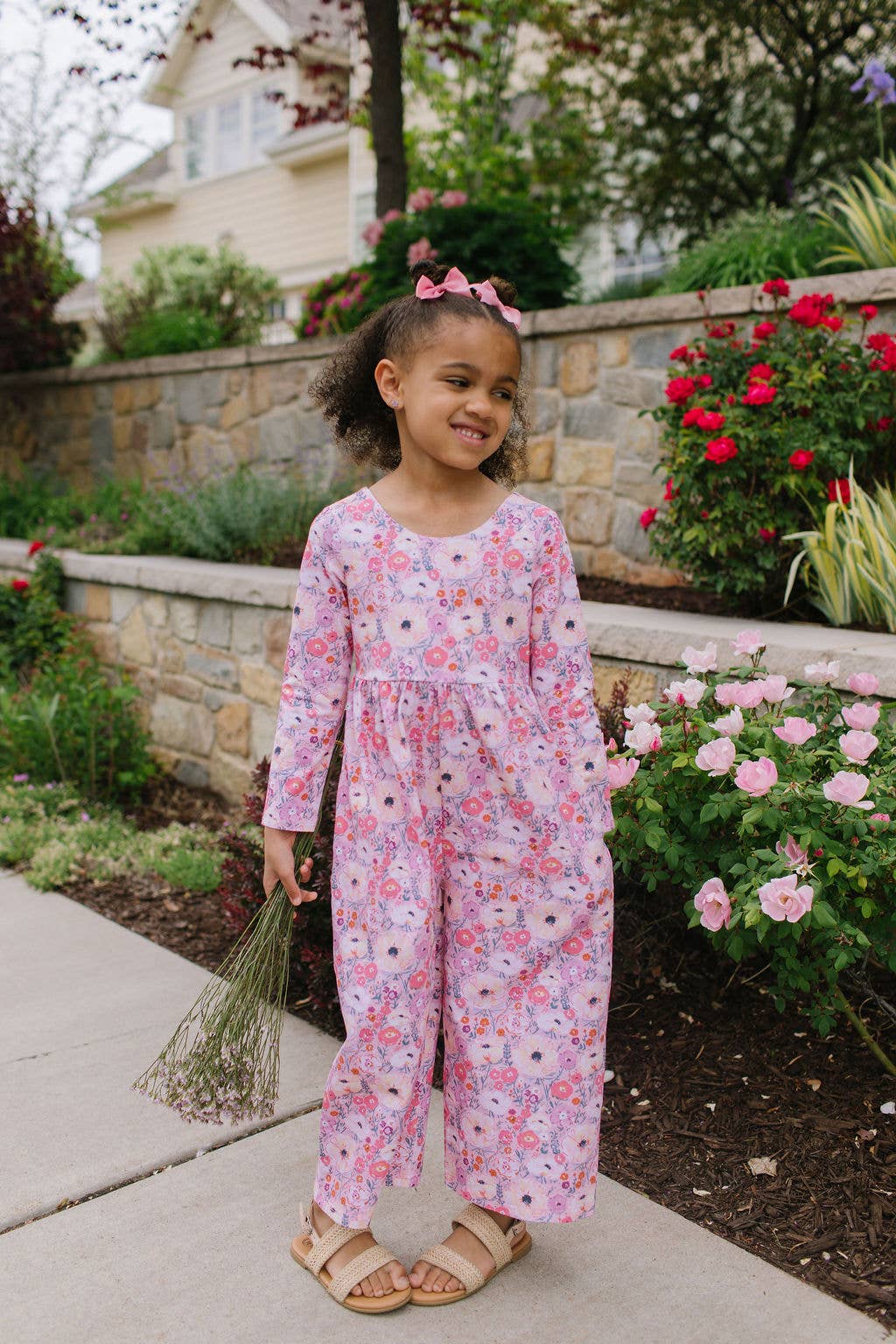 Young girl in a floral outfit standing outdoors with flowers and a garden in the background