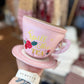 Pink teacup-shaped pouch with floral design and text on a blurred background