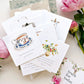 Stack of greeting cards with floral and teacup designs on a light surface with flowers.