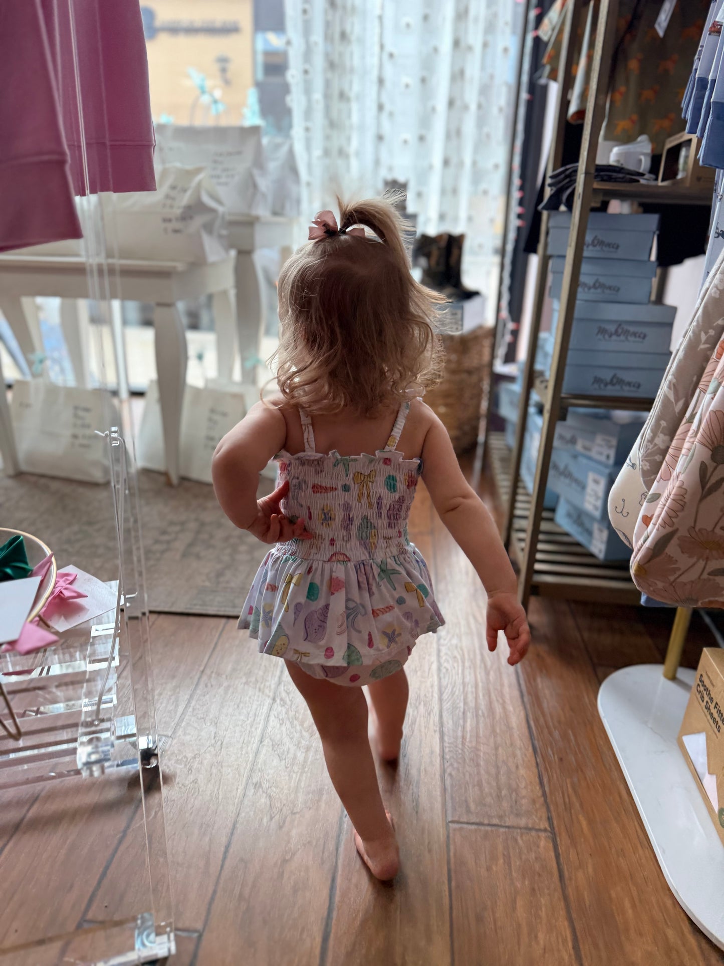 Child in a patterned dress standing in a room with wooden floor and furniture.