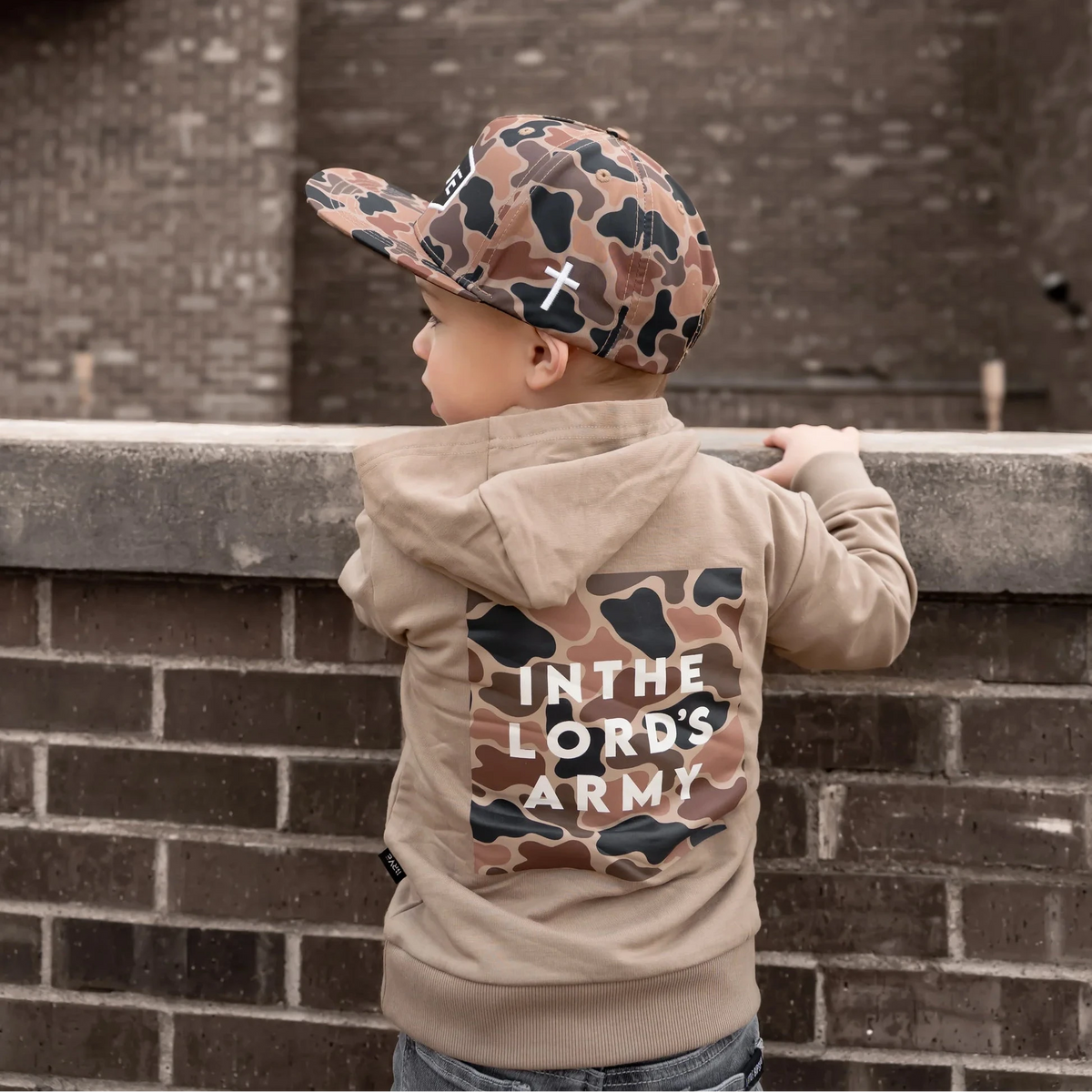 Child wearing a hoodie with 'In the Lord's Army' text, standing against a brick wall.