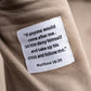 A close-up of a beige fabric garment with a white rectangular patch sewn onto it. The patch contains a black printed Bible verse from Matthew 16:24: “If anyone would come after me, let him deny himself and take up his cross and follow me.