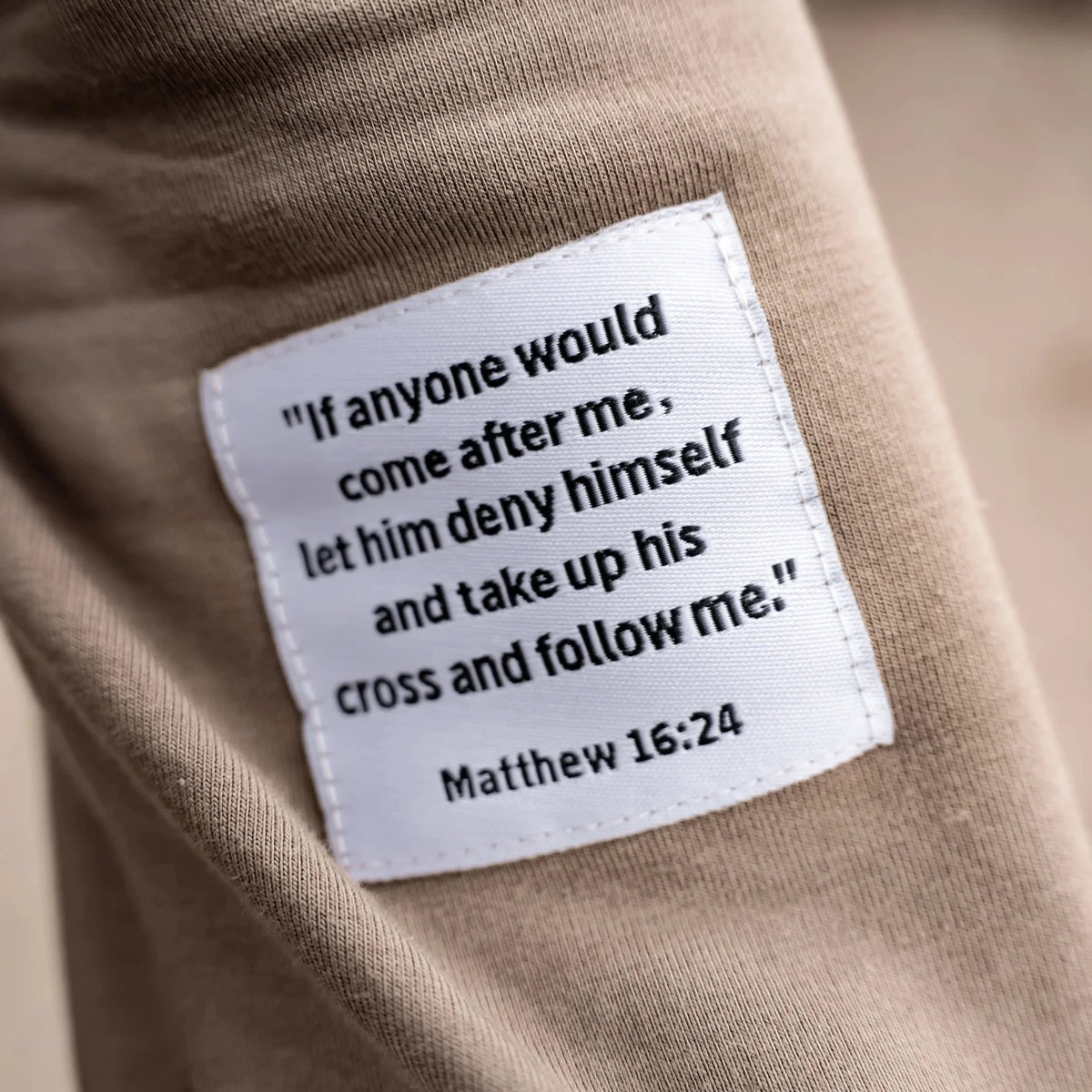 A close-up of a beige fabric garment with a white rectangular patch sewn onto it. The patch contains a black printed Bible verse from Matthew 16:24: “If anyone would come after me, let him deny himself and take up his cross and follow me.