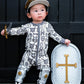A toddler wearing a medieval knight costume stands in front of a dark wooden door, holding a toy sword and a toy shield. The costume includes a white and gray printed jumpsuit with knight motifs, a gold helmet, and silver shin guards.