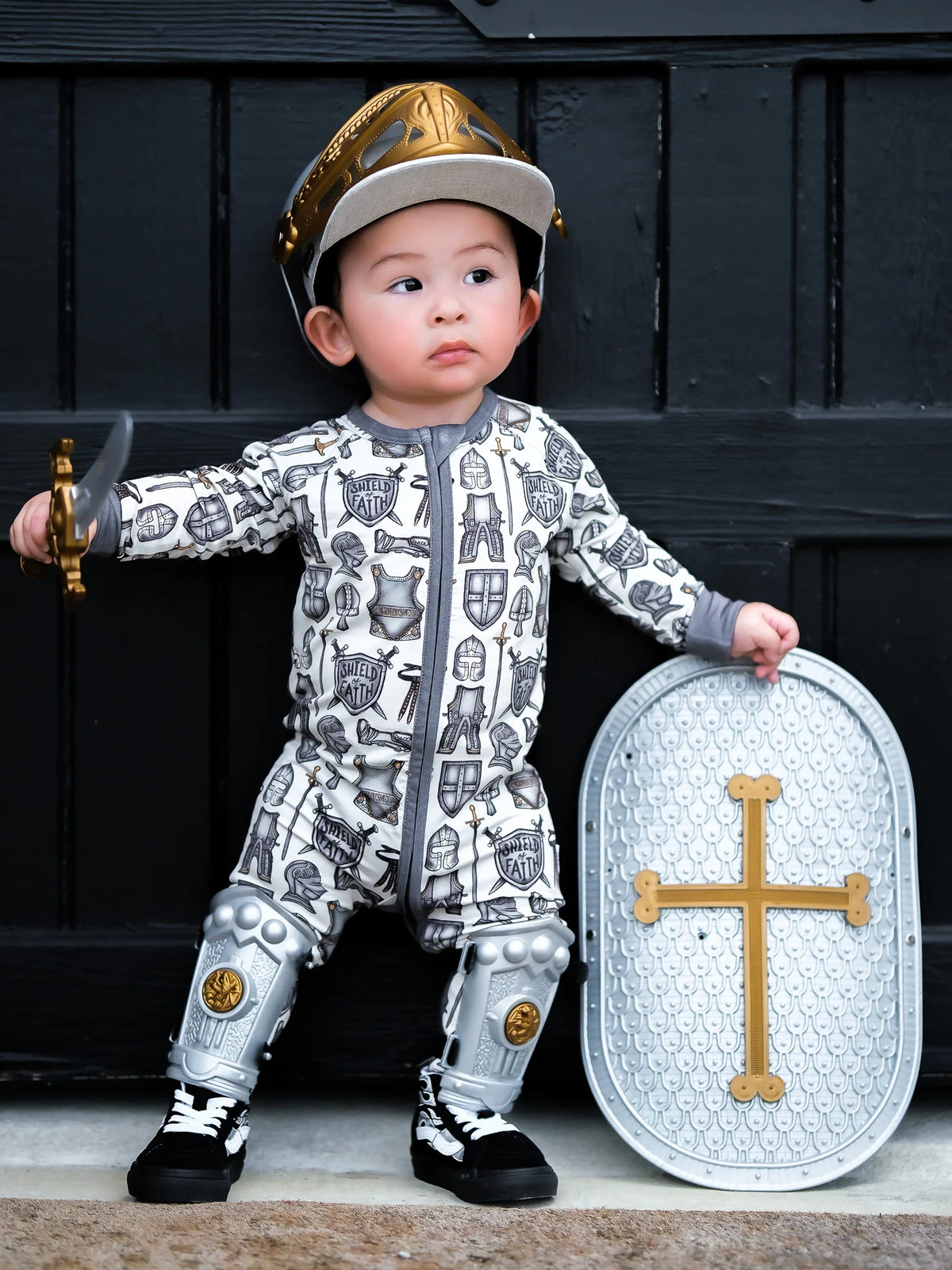 A toddler wearing a medieval knight costume stands in front of a dark wooden door, holding a toy sword and a toy shield. The costume includes a white and gray printed jumpsuit with knight motifs, a gold helmet, and silver shin guards.