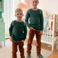 Two children in matching green sweaters and brown pants standing in a room with a crib and dresser.
