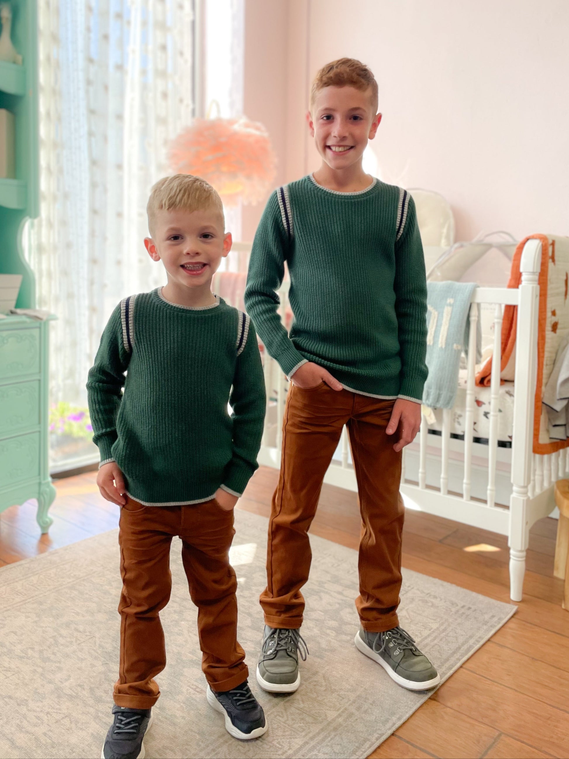 Two children in matching green sweaters and brown pants standing in a room with a crib and dresser.