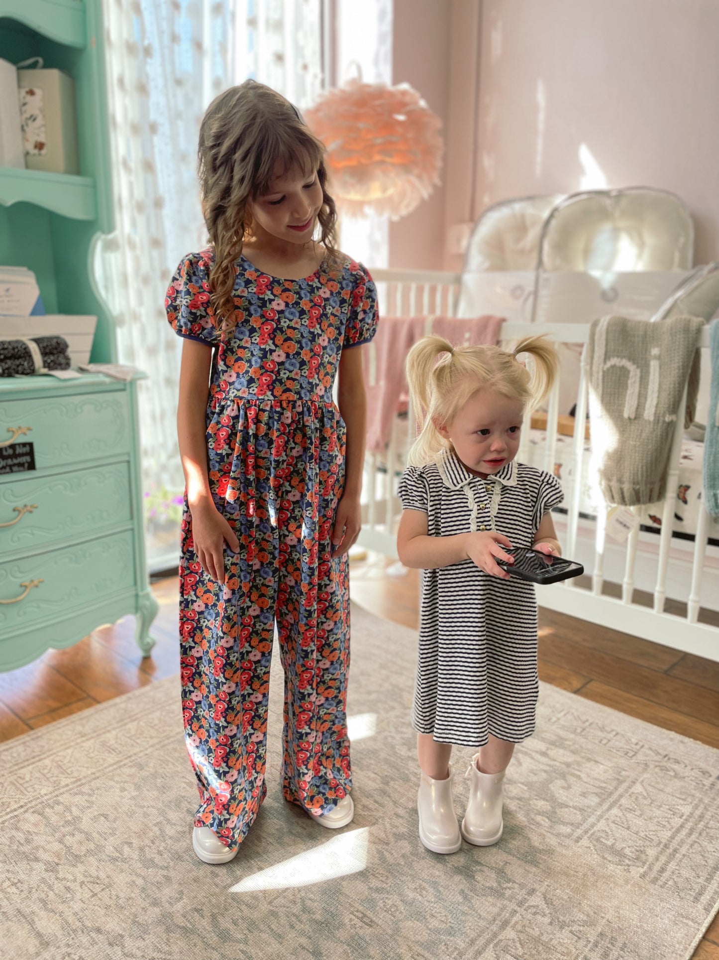 Two young girls in a nursery, one in a floral jumpsuit and the other in a striped dress, standing on a rug.