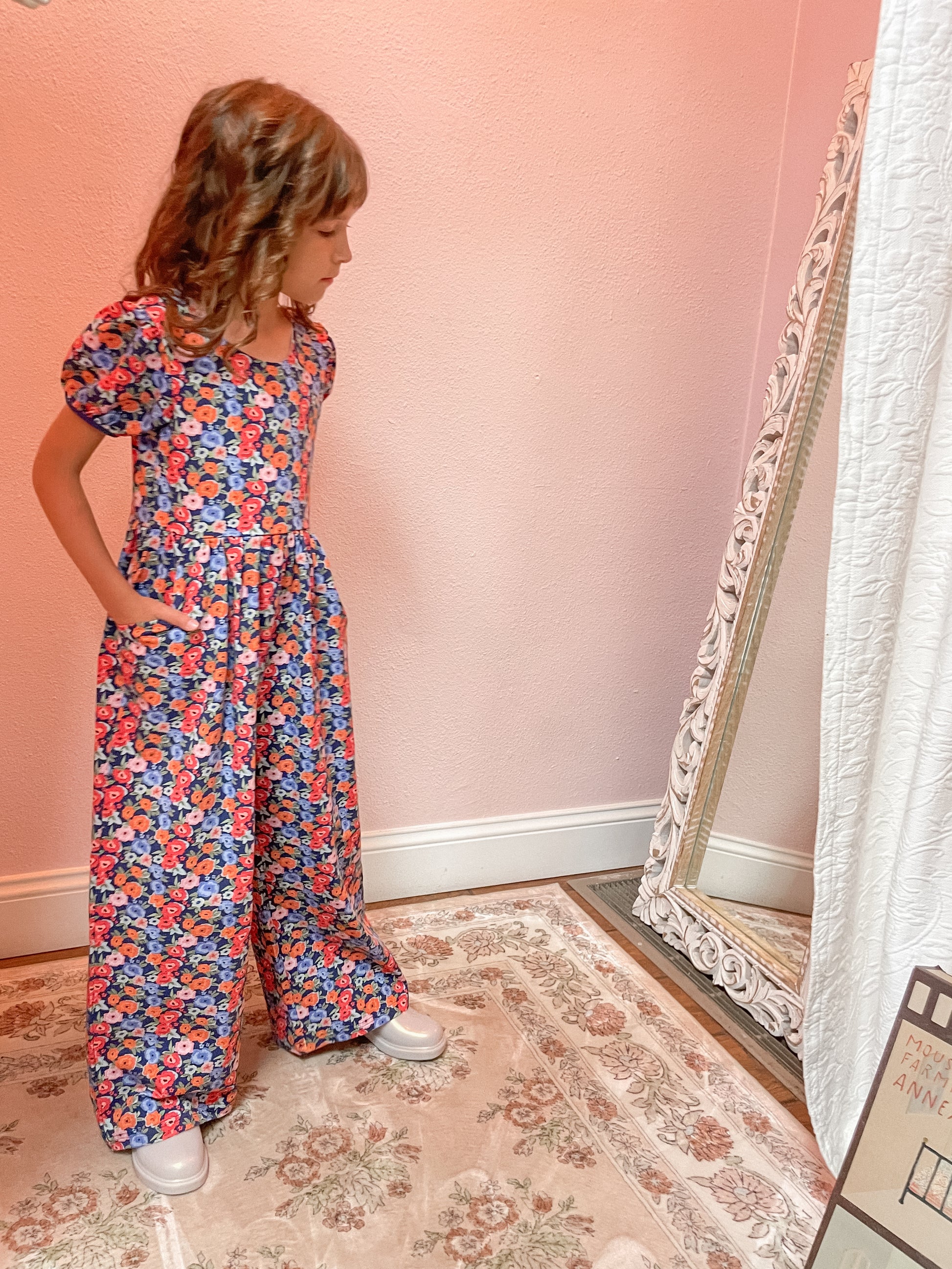Young girl in a floral romper standing in front of a mirror on a patterned rug.
