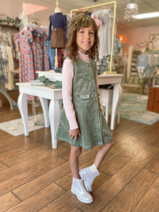 Young girl in a green dress standing in a store with wooden floors and clothing racks in the background.