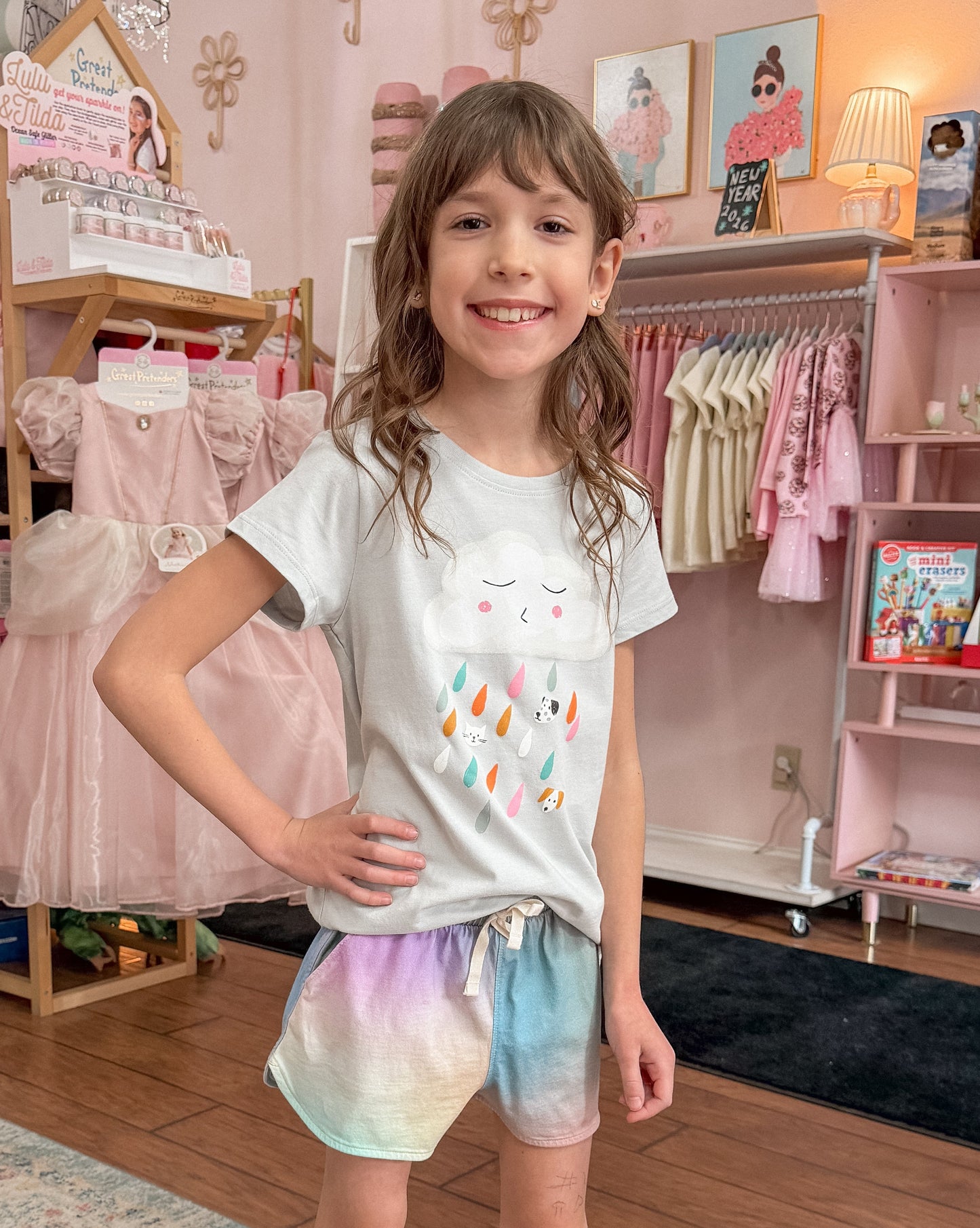 Young girl in a clothing store wearing a light grey t-shirt with a cloud graphic raining colorful raindrops, and colorful watercolor looking shorts.