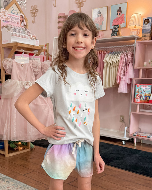 Young girl in a clothing store wearing a light grey t-shirt with a cloud graphic raining colorful raindrops, and colorful watercolor looking shorts.