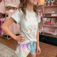 Young girl wearing a white t-shirt with colorful designs and pastel shorts in a store setting.