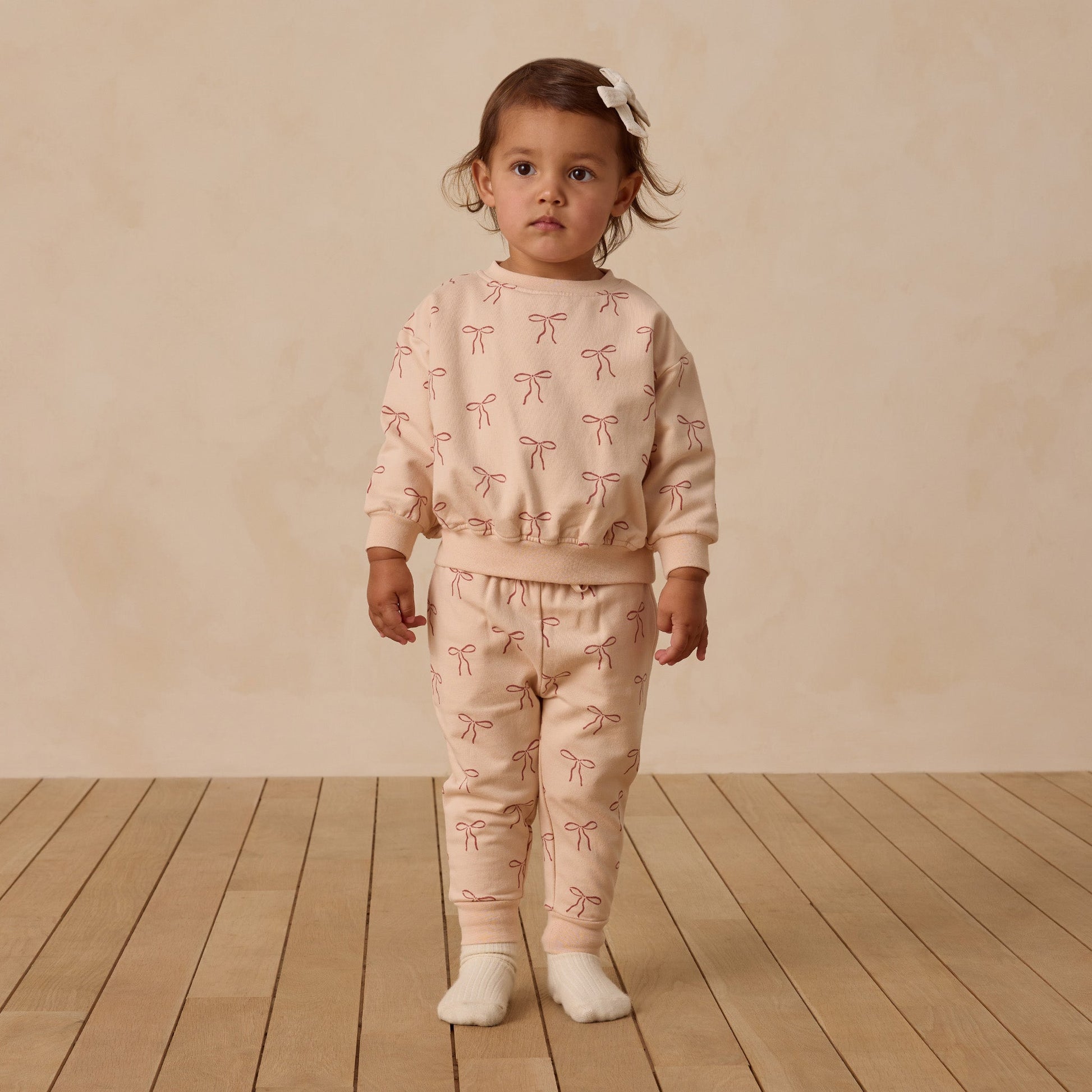 Child wearing a pink outfit with bow patterns on a wooden floor and beige wall background
