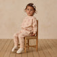 Young child wearing a pink outfit with bow patterns, sitting on a wooden chair against a beige wall.