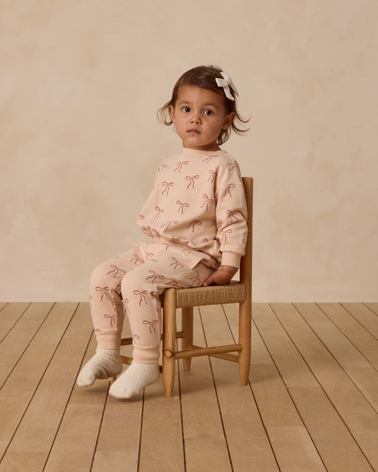 Young child wearing a pink outfit with bow patterns, sitting on a wooden chair against a beige wall.