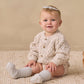 A baby sitting on a textured cream rug wearing a cream cable-knit cardigan, matching knit socks, and a small white bow headband, with a wicker basket nearby against a neutral beige backdrop.