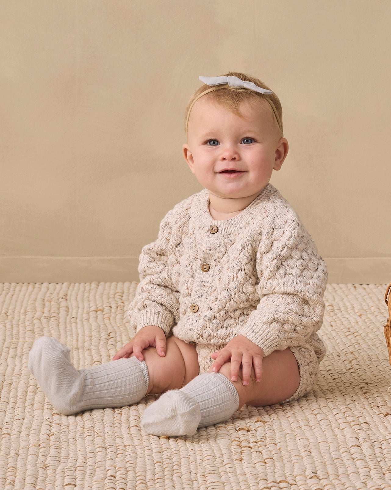 A baby sitting on a textured cream rug wearing a cream cable-knit cardigan, matching knit socks, and a small white bow headband, with a wicker basket nearby against a neutral beige backdrop.