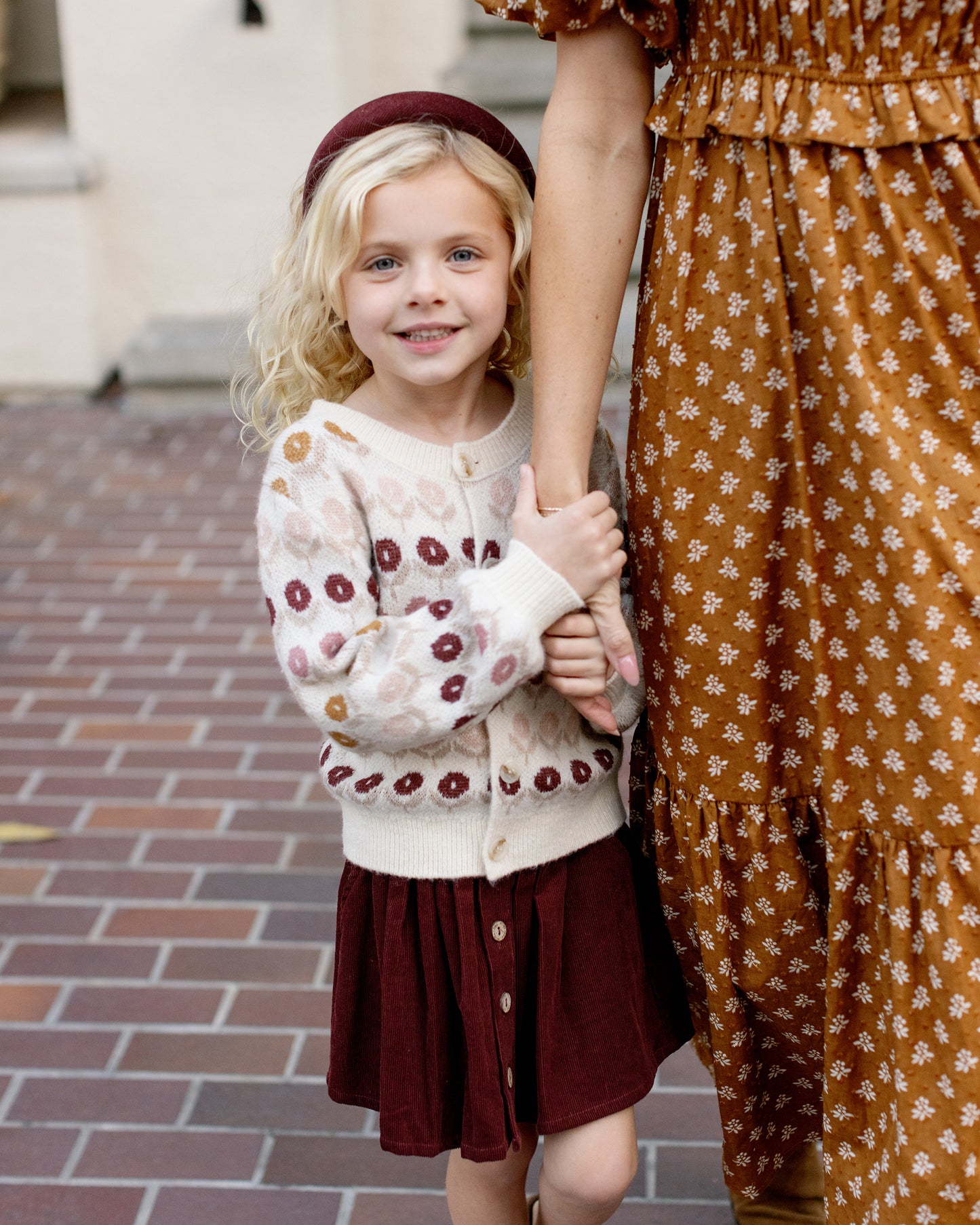 Young girl in a patterned sweater and skirt holding hands with an adult on a brick path.