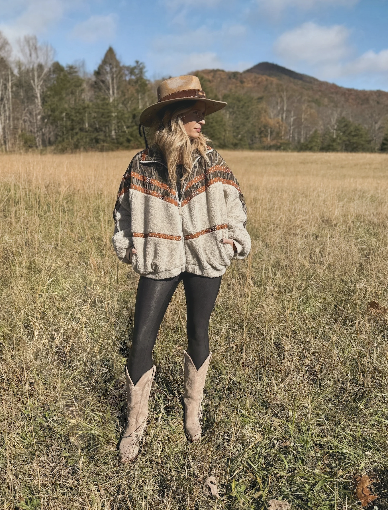Person wearing a patterned sweater, hat, and boots standing in a field with mountains in the background
