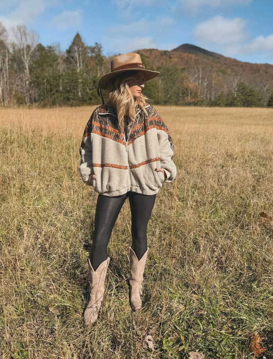 Person wearing a patterned sweater, hat, and boots standing in a field with mountains in the background