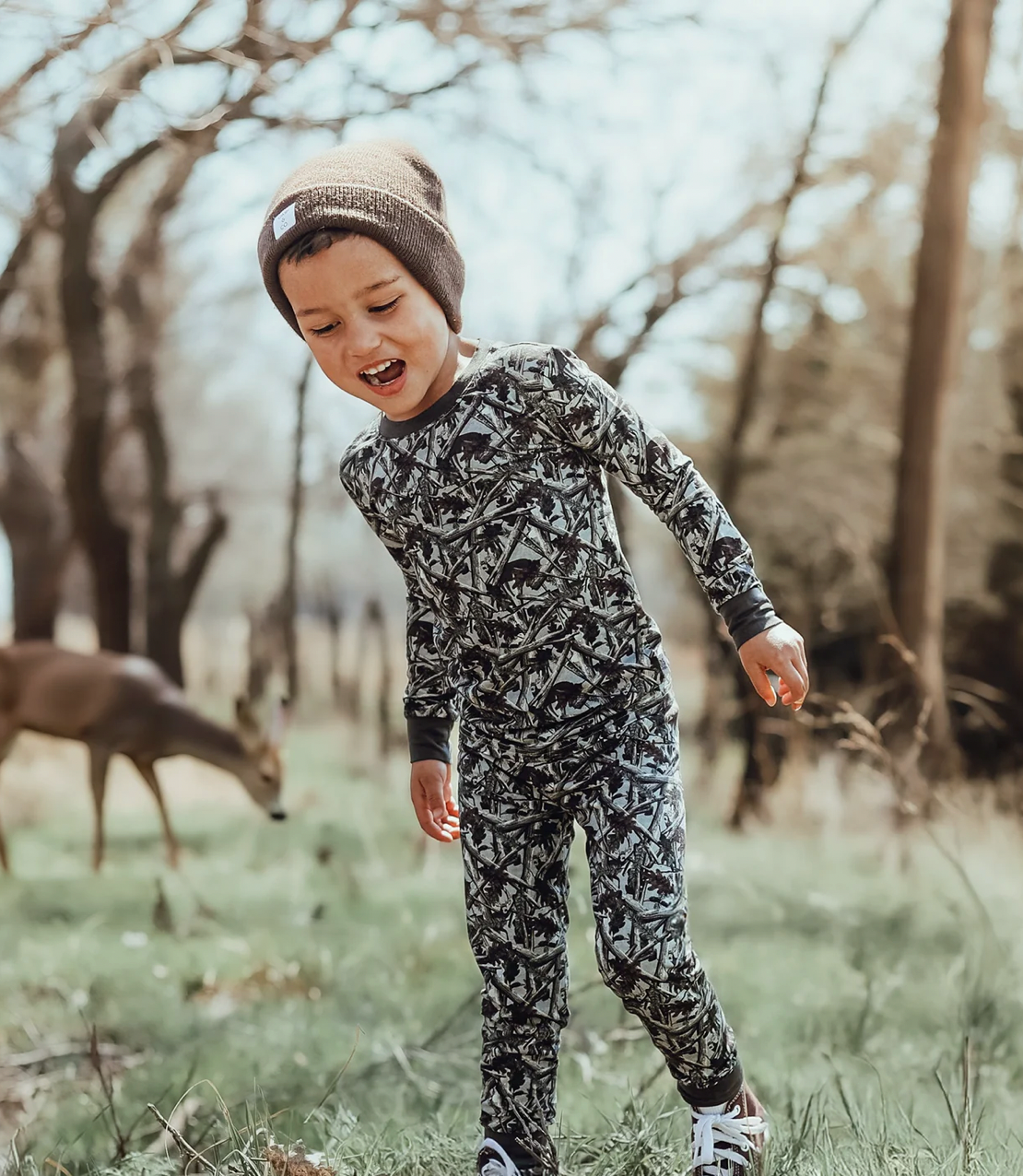 Child in camouflage outfit standing in a forest with deer in the background