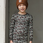 Child wearing a camouflage long-sleeve shirt and pants with a brown beanie, standing against a neutral background.
