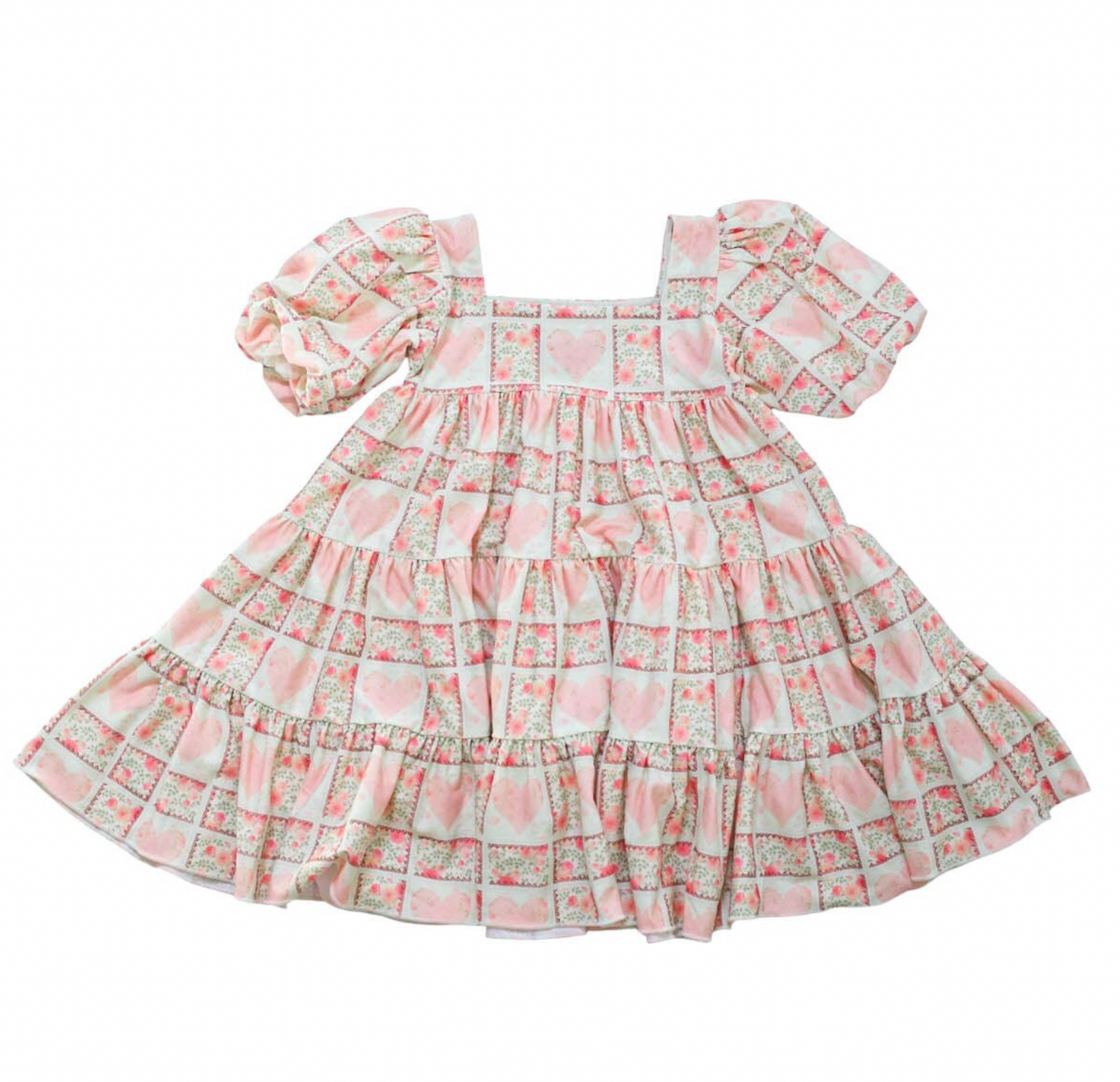 Puff sleeve dress on white background, dress has a block print reminiscent of quilting with faint watercolor looking pink hearts in the center on some blocks, and a floral print in other blocks. The skirt of the dress is sewn into three separate tiers, and the waist is in the empire style with a square neckline. 