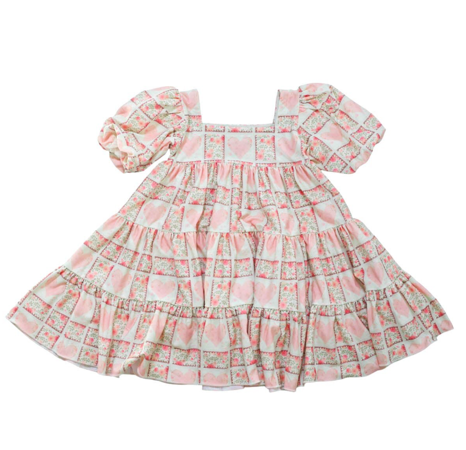 Puff sleeve dress on white background, dress has a block print reminiscent of quilting with faint watercolor looking pink hearts in the center on some blocks, and a floral print in other blocks. The skirt of the dress is sewn into three separate tiers, and the waist is in the empire style with a square neckline. 