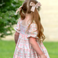 little girl playing in an outdoor scene with lots of greenery, wearing Puff sleeve dress with a soft white background, dress has a block print reminiscent of quilting with faint watercolor looking pink hearts in the center on some blocks, and a floral print in other blocks. The skirt of the dress is sewn into three separate tiers, and the waist is in the empire style with a square neckline. 