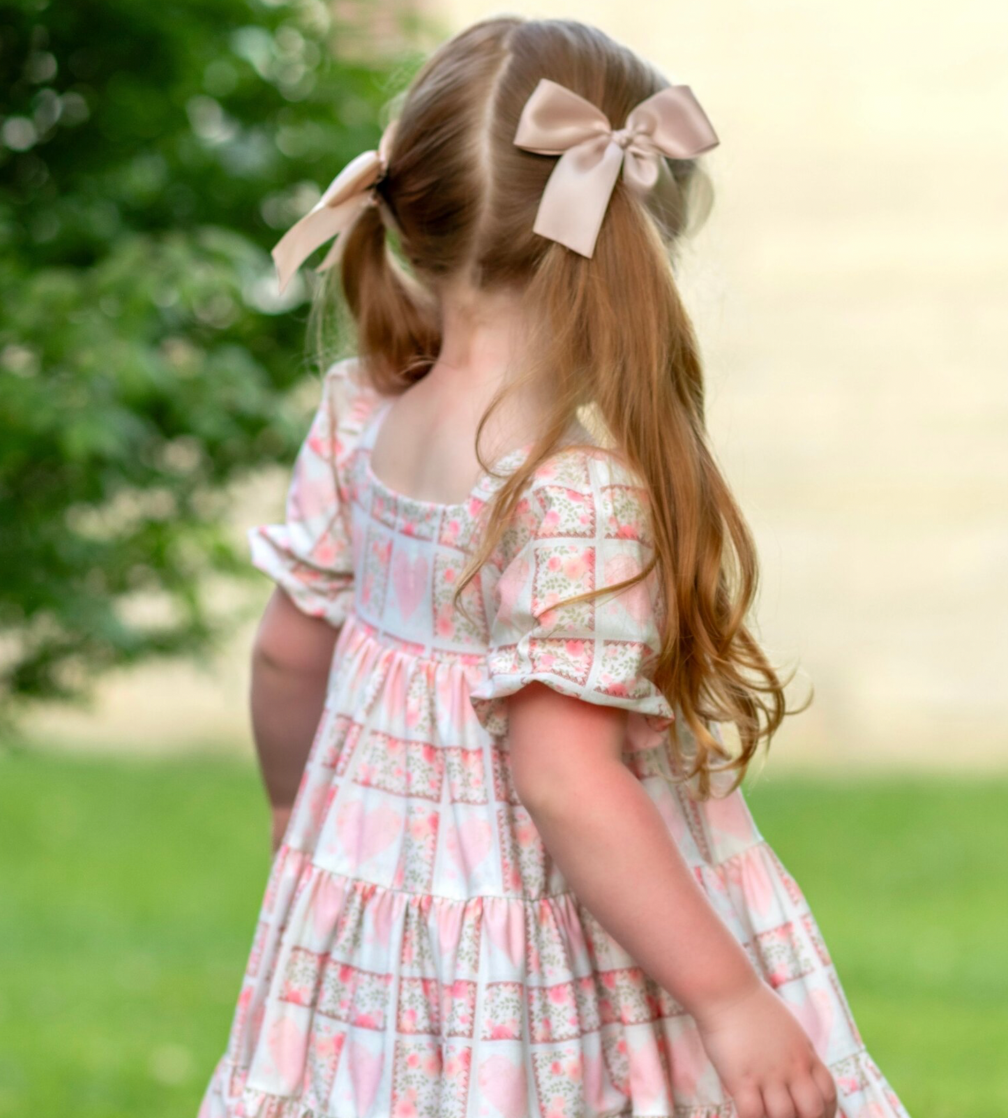 little girl playing in an outdoor scene with lots of greenery, wearing Puff sleeve dress with a soft white background, dress has a block print reminiscent of quilting with faint watercolor looking pink hearts in the center on some blocks, and a floral print in other blocks. The skirt of the dress is sewn into three separate tiers, and the waist is in the empire style with a square neckline. 