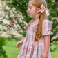 little girl playing in an outdoor scene with lots of greenery, wearing Puff sleeve dress with a soft white background, dress has a block print reminiscent of quilting with faint watercolor looking pink hearts in the center on some blocks, and a floral print in other blocks. The skirt of the dress is sewn into three separate tiers, and the waist is in the empire style with a square neckline. 